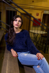 Fototapeta premium A woman in a blue sweater and jeans sits on stage steps, looking thoughtful and composed.