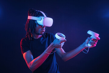 White controllers in hands, playing game. African American guy is with virtual reality glasses in...
