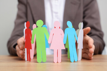 Woman protecting paper human figures at wooden table, closeup. Diversity and inclusion concept