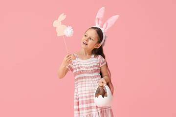 Cute little girl in bunny ears holding basket with sweet chocolate egg and paper rabbit on pink background. Easter celebration
