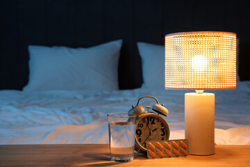 Alarm clock with melatonin pills, glass of water and glowing lamp on table in bedroom at night