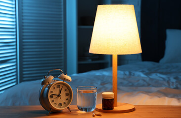 Alarm clock with melatonin pills, glass of water and glowing lamp on table in bedroom at night