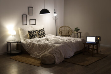 Interior of modern bedroom with cozy bed, laptop and glowing lamps at night
