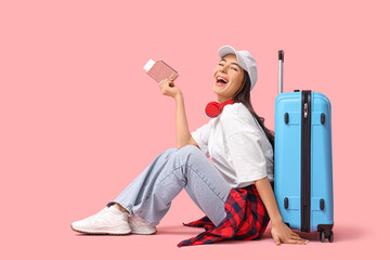Happy young female tourist with passport and suitcase sitting on pink background