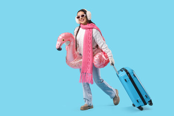 Young female tourist in winter clothes with suitcase and inflatable ring walking on blue background