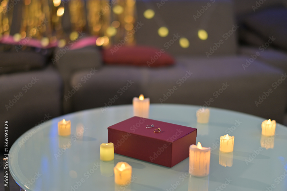 Poster romantic setup with red gift box and rings on glass table surrounded by glowing candles, creating wa