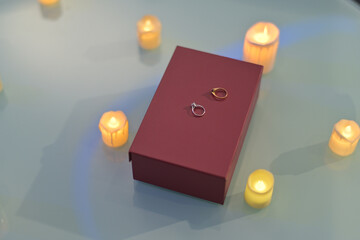 red gift box with two elegant rings on top, surrounded by glowing candles on glass surface, creating romantic ambiance