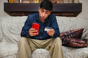 Upset Indianian man sit on sofa at home holding credit card looking at mobile phone screen, showing...