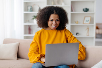 Young African American woman working on laptop while sitting on couch at home, millennial black...