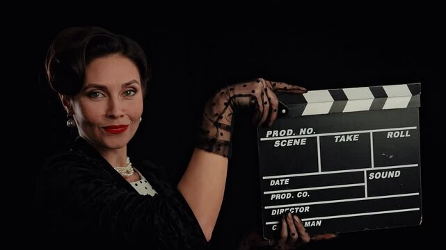 Retro woman actress with pin-up hairstyle and makeup isolated on black background, turns at camera and claps the clapperboard. Slow motion.