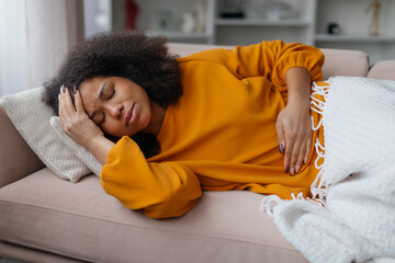 Sick black woman lying on couch, feeling unwell at home, ill african american female suffering from...