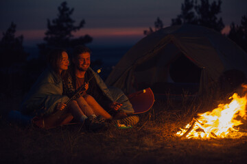 Young tourist couple exploring new places. An attractive woman and a handsome man spend time together in nature. Sitting by the campfire and tent in the evening mountains and making marshmallows. © dsheremeta