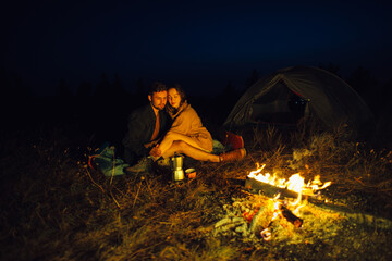 A happy couple of tourists hugs while warming themselves by a campfire against the background of a tent and night mountains. The concept of hiking in nature.