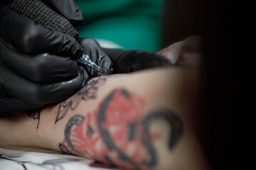 Close up tattoo master professional tattoo artist in black gloves making a rose tattoo on a man's forearm. He is holding a tattoo machine.