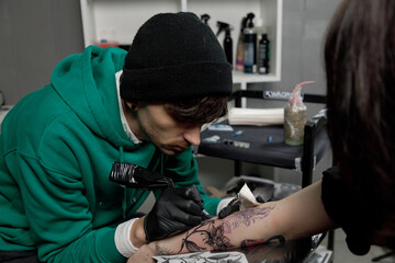 Close up tattoo master professional tattoo artist in black gloves making a rose tattoo on a man's forearm. He is holding a tattoo machine.
