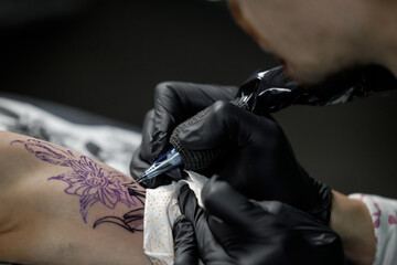 Close up tattoo master professional tattoo artist in black gloves making a rose tattoo on a man's forearm. He is holding a tattoo machine.