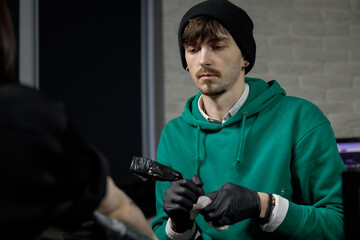 Close up tattoo master professional tattoo artist in black gloves making a rose tattoo on a man's forearm. He is holding a tattoo machine.