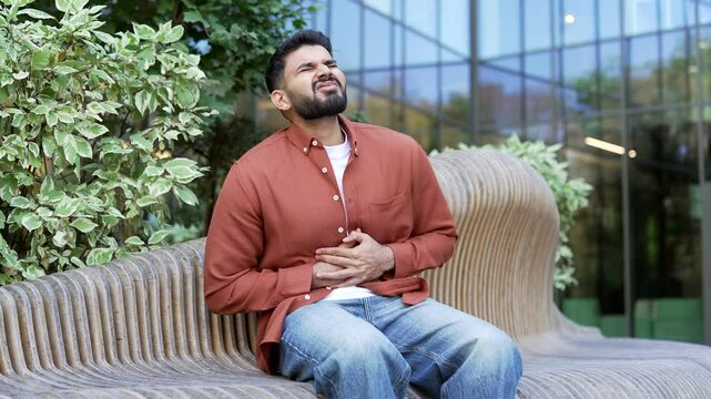 Businessman feels stomach pain while sitting on a bench on the street near business office building. Sick man has heartburn, gastritis, bloating or poisoning. Male suffers from spasms and constipation