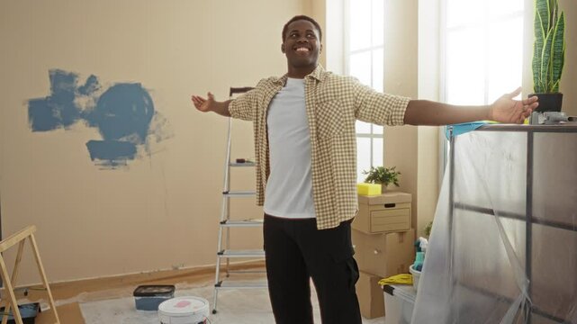 Young man indoors smiling with arms outstretched in a newly decorated room with paint supplies scattered around and a ladder against the wall.