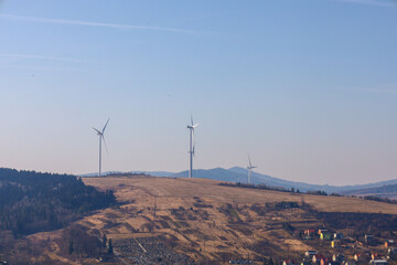 The photograph depicts a panoramic view of a hill with wind turbines located on its top. In the foreground, a part of the city with colorful houses and a cemetery is visible.