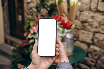 Mockup woman's hand holding using smart phone with white blank screen POV at flower street shop, mobile app design and advertising, online marketing