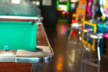 Cue chalk near chrome cast corners of classic pool tables with blurry colorful illuminated arcade game machines background at indoor game room in Houston, Texas, green playfield cloth covered