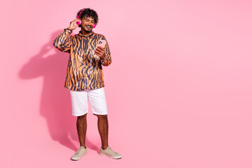 Young stylish man with curly hair using smartphone while listening to music wearing trendy outfit against vibrant pink background