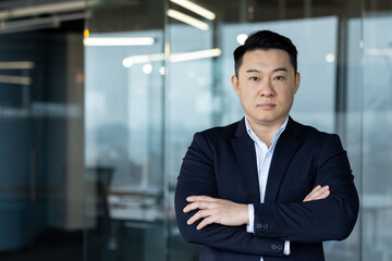 Fototapeta premium Portrait of a young serious Asian businessman in a business suit standing in the office, arms crossed on his chest and looking confidently at the camera