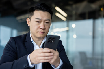 Close-up photo of a young focused and serious Asian businessman sitting in the office and using his...