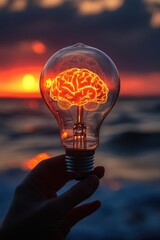 Hand holding a light bulb with a glowing brain with a blurry sunset background