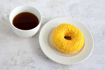 Yellow glazed donut with sprinkles on a white plate next to a cup of coffee