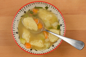 A top-down shot presents a bowl of chicken soup with semolina dumplings, carrots, and greens, with a spoon resting inside