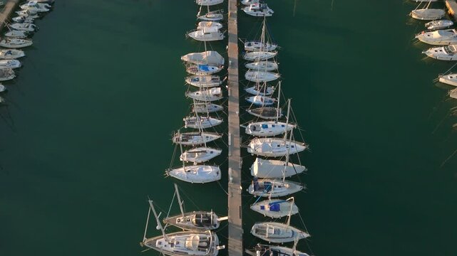 Top-down drone view of multiple sailboats moored neatly along a central pier in a calm marina. 