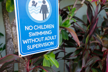 Blue safety sign stating "NO CHILDREN SWIMMING WITHOUT ADULT SUPERVISION" mounted among tropical plants