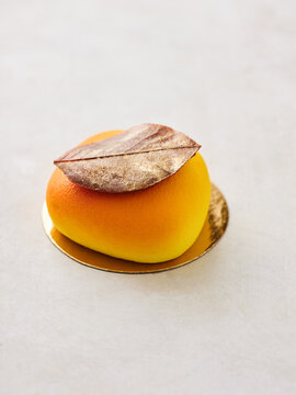 An elegant dessert shaped like a fruit sits on a gold plate, adorned with a metallic leaf. The vibrant colors of yellow and gold create a stunning presentation ideal for special occasions