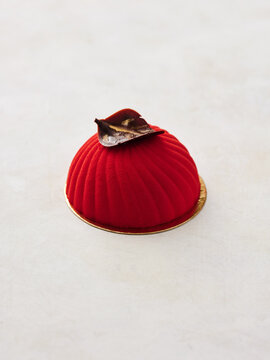An exquisite red dessert sits atop a golden base, showcasing smooth curves and a striking chocolate leaf garnish. The minimalist setting enhances its vibrant color and elegant design