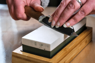 A close-up of a person sharpening a knife on a whetstone. The hands guide the blade at the correct...