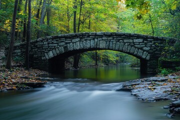 Fototapeta premium Old stone bridge arches gracefully over a flowing river through a forest. Great for travel brochures or highlighting natural beauty in promotions.