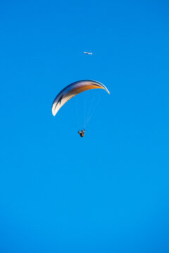 A paraglider floats in the clear blue sky with a distant aircraft above and in the background.