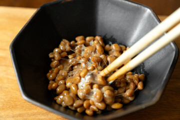 箸でかき混ぜてネバネバの糸を引いている納豆　黒いボウル　木製テーブルの背景