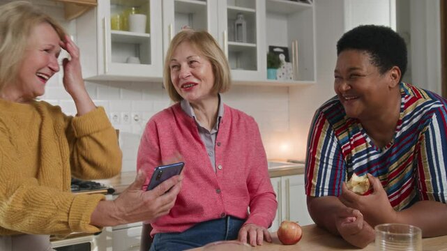 Senior Women Friends Gathering at Home. Three cheerful 60 year old women enjoying get together in living room and chatting. Leisure time Three mature women using and looking in mobile telephone.