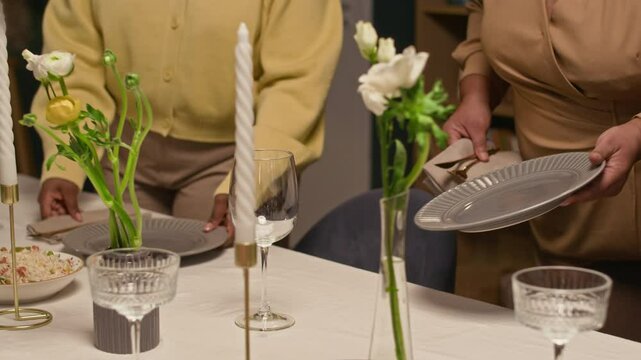 Cropped shot of Black women placing empty plates on dining table decorated with candles while setting up festive dinner together during family celebration at home party, dolly away motion
