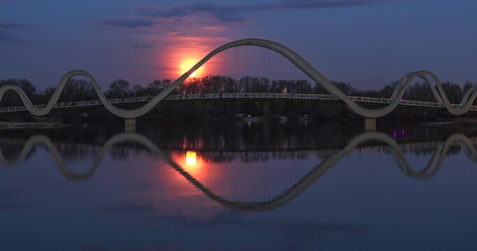 Full moonrise over a pedestrian bridge in Obolonskyi District, Kyiv, Ukraine, reflecting in the calm waters, creating a mesmerizing symmetrical scene at dusk. Time lapse
