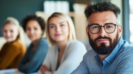 A focused man with a beard sits in a meeting room, surrounded by three women, all engaged in a discussion.