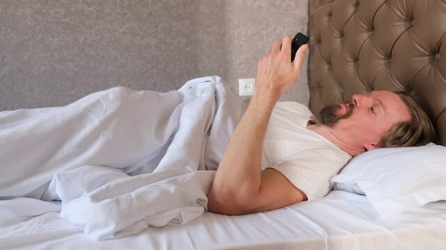 Young attractive man uses smartphone to watch videos and masturbates under the blanket in bed satisfying his sexual desire on his own. Porn addiction and excess hormone.