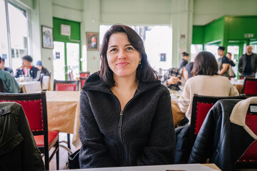 A smiling woman in a black fleece jacket sits at a table in a busy restaurant with green walls, surrounded by other diners enjoying their meals.. © Raivo