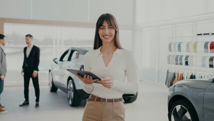 Busy Caucasian female standing in middle of car dealership. Using tablet device with large display....