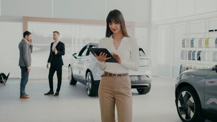 Beautiful Caucasian woman typing on big screen of tablet device. Manager working at car dealership. Managing operations online. Looking at camera and smiling. Bright showroom. Modern automobiles.