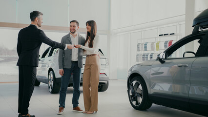 Couple standing in bright auto showroom. Woman widely smiling while car dealer handing her car...