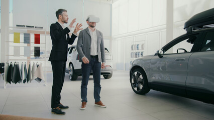 Man in grey jacket using VR headset. Worker standing nearby and guiding or explaining experience. Spacious room with cars and material samples. Showroom or automotive studio. Exploring new car models.